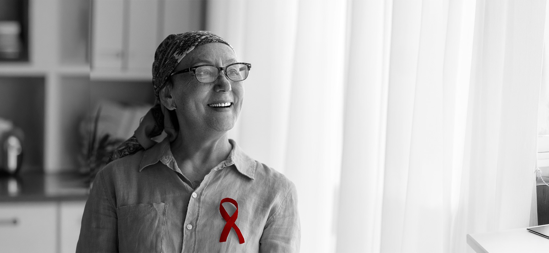 A imagem mostra uma mulher de meia-idade, com câncer, usando lenço na cabeça, enquanto sorri. Além de um broche simbolizando a luta contra o câncer.