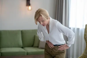 A imagem mostra uma mulher de meia-idade, em sua casa, de pé. apoiando a barriga com as mãos, sentindo dor. Ilustra o texto sobre Doença de Crohn .