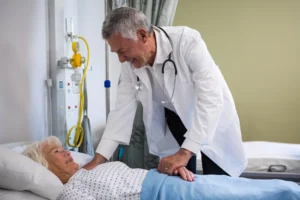 A imagem mostra um médico sênior, usando jaleco e estetoscópio em volta do pescoço, atendendo uma paciente que está acamada. Ambos sorriem. Ilustra o texto sobre a aposentadoria especial do médico servidor público.