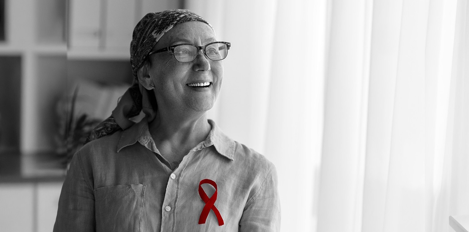 A imagem mostra uma mulher de meia-idade, com câncer, usando lenço na cabeça, enquanto sorri. Além de um broche simbolizando a luta contra o câncer.