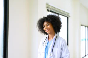 A imagem mostra uma jovem médica em ambiente hospitalar, sorrindo, usando um jaleco e estetoscópio em volta do pescoço. Ilustra o texto sobre residência médica e aposentadoria.