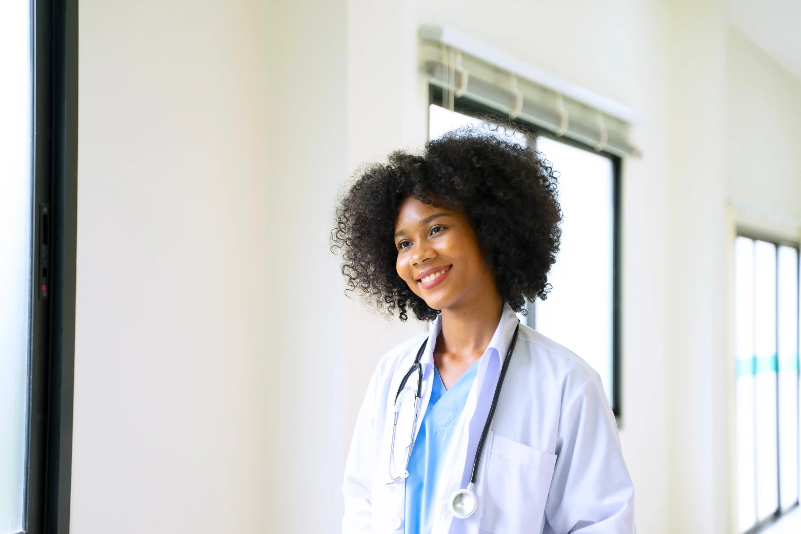 A imagem mostra uma jovem médica em ambiente hospitalar, sorrindo, usando um jaleco e estetoscópio em volta do pescoço. Ilustra o texto sobre residência médica e aposentadoria.