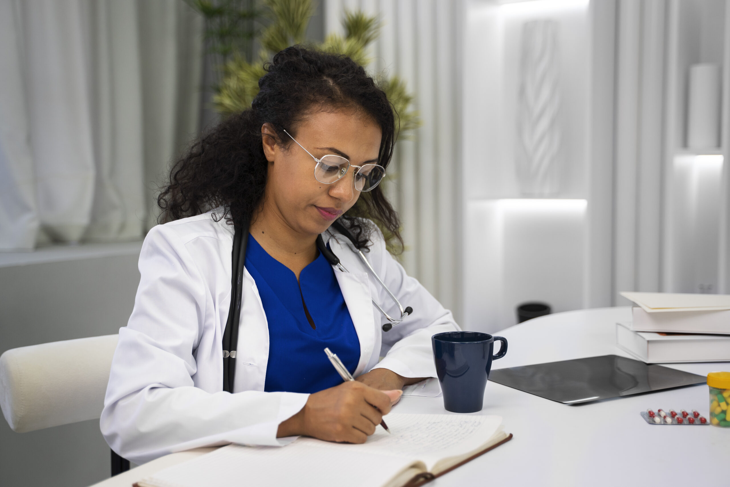 Médica sentada em consultório escrevendo em caderno, usando jaleco branco e estetoscópio, com mesa de trabalho, xícara e materiais médicos ao redor.