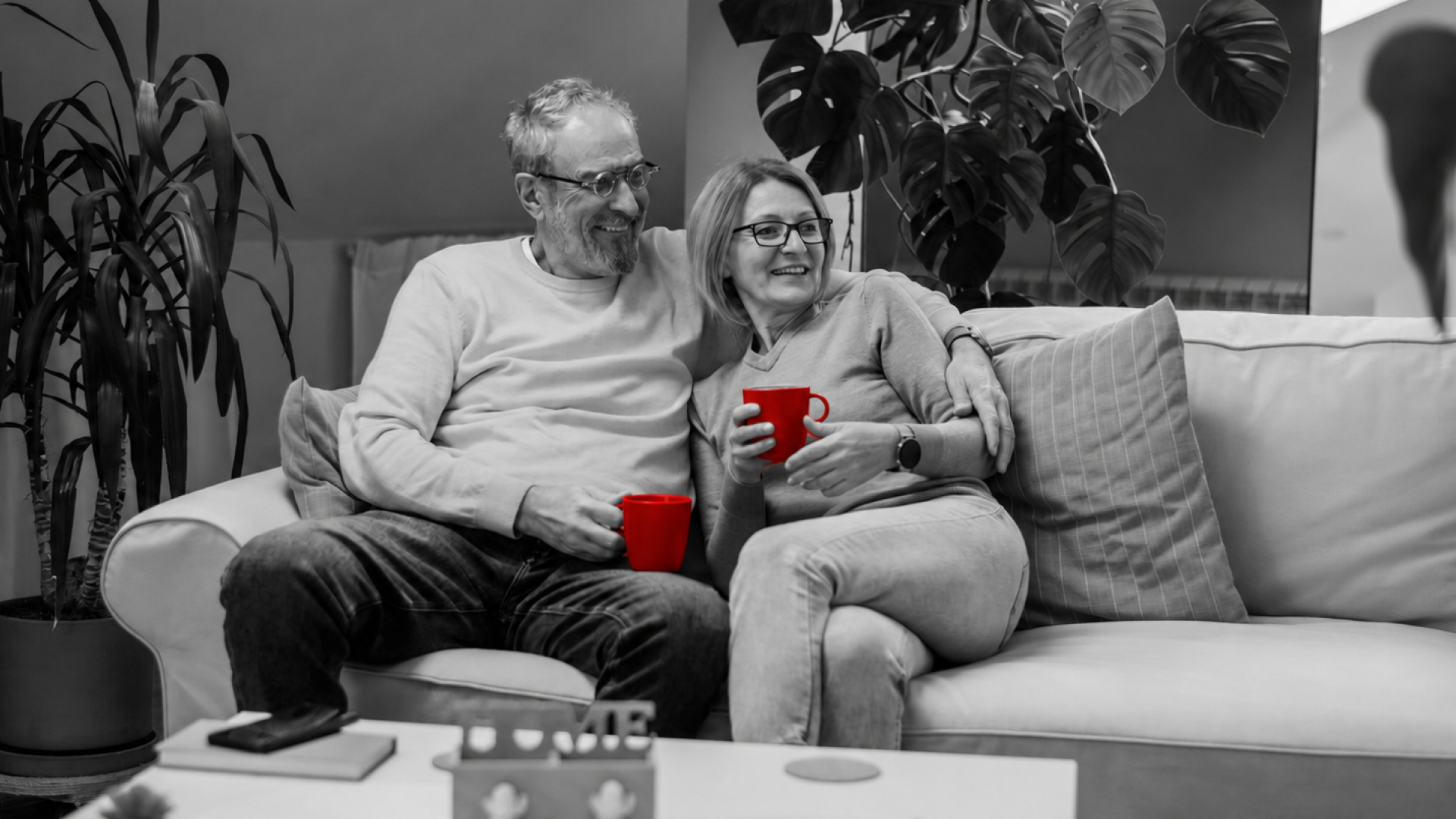 A imagem mostra um casal de meia-idade, em casa, ambos sentados um sofá com xícaras de café em uma das mãos.