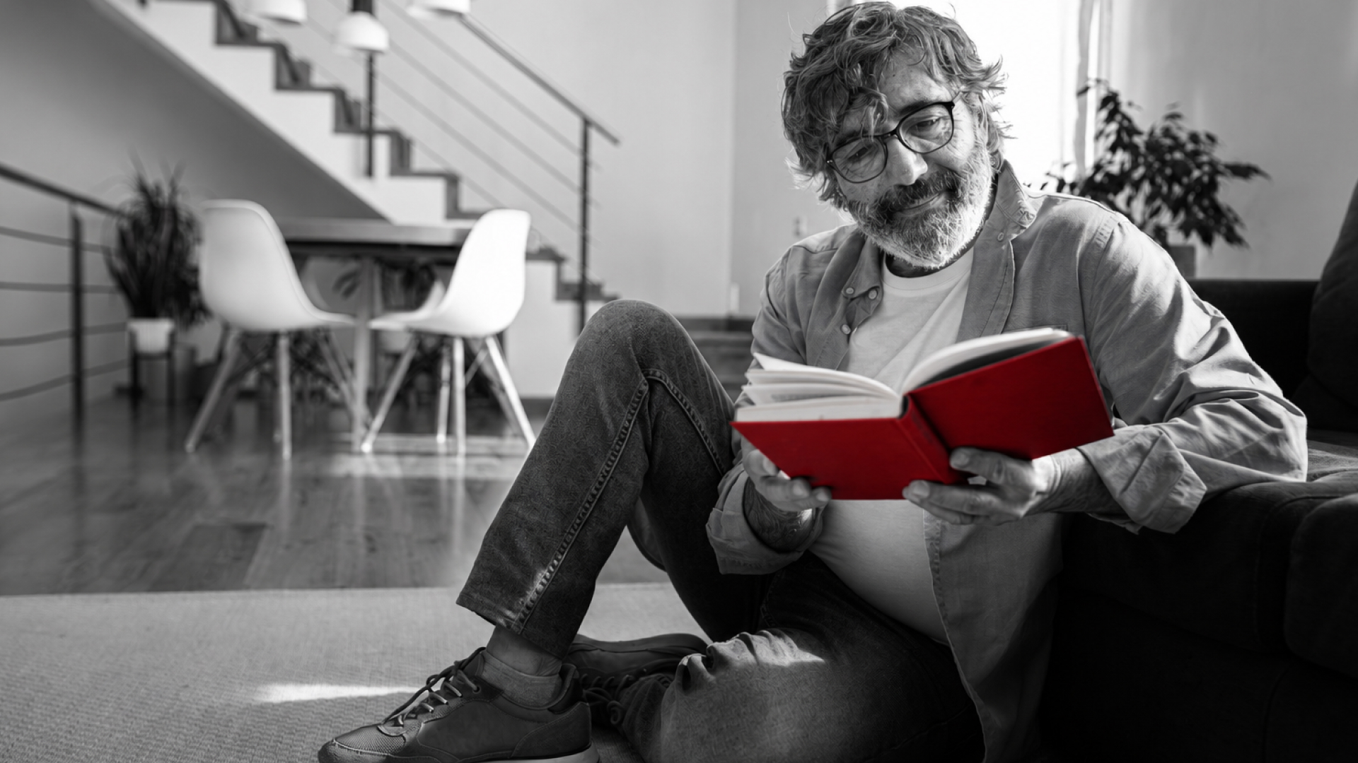A imagem mostra um homem de meia-idade, usando óculos de grau, sentado no chão de sua casa, lendo um livro.