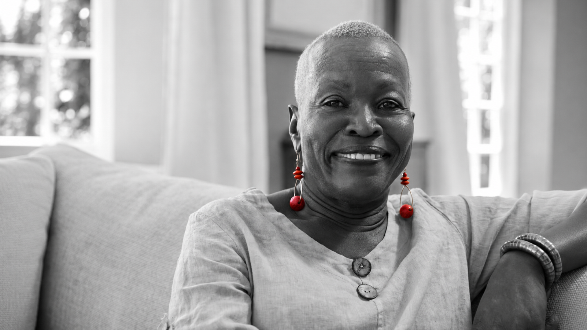 A imagem mostra uma mulher de meia-idade, sentada em seu sofá, dentro de casa, sorrindo para a câmera.