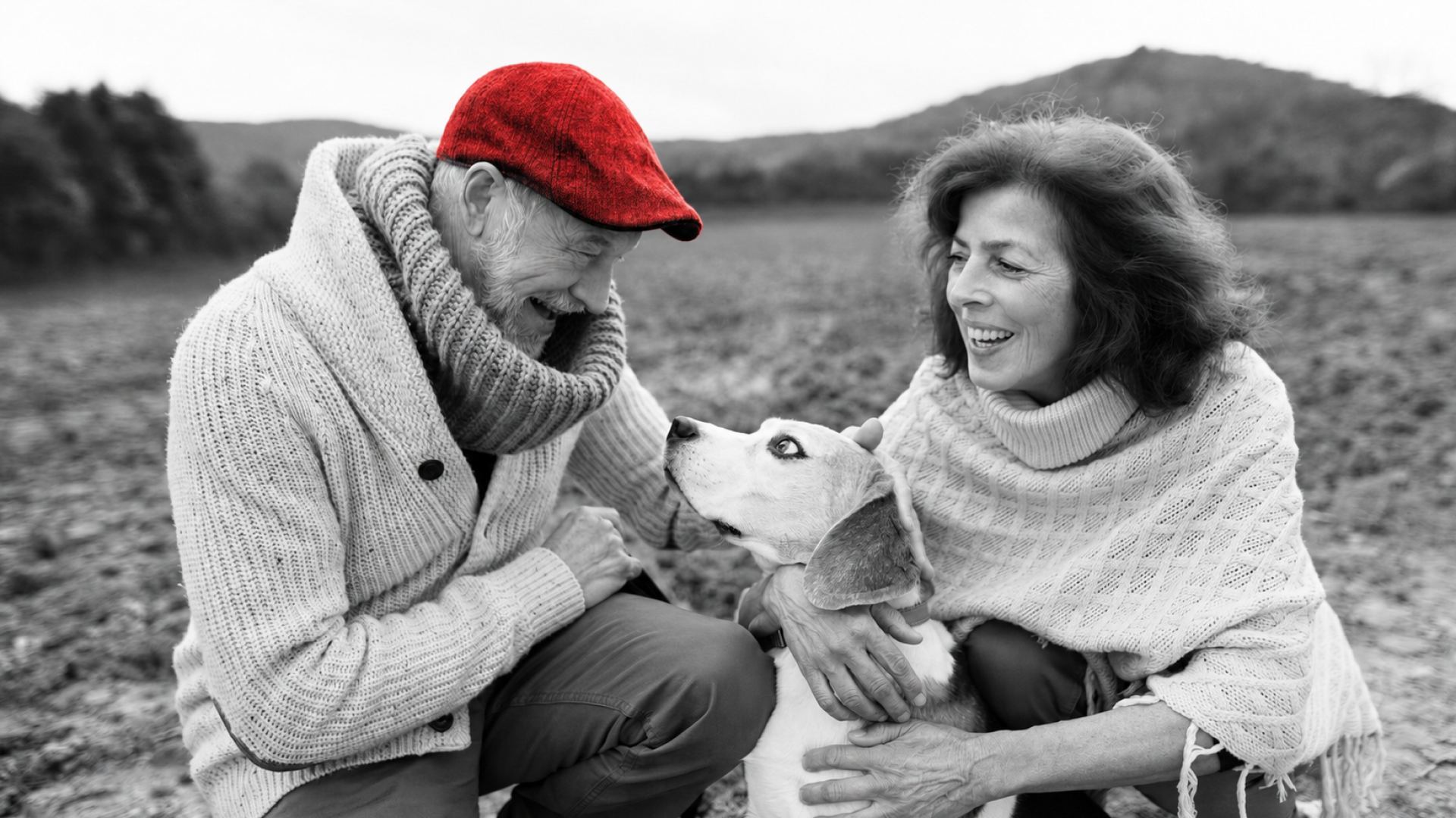 A imagem mostra um casal de meia-idade, sorrindo, ao ar livre, com seu cachorro.