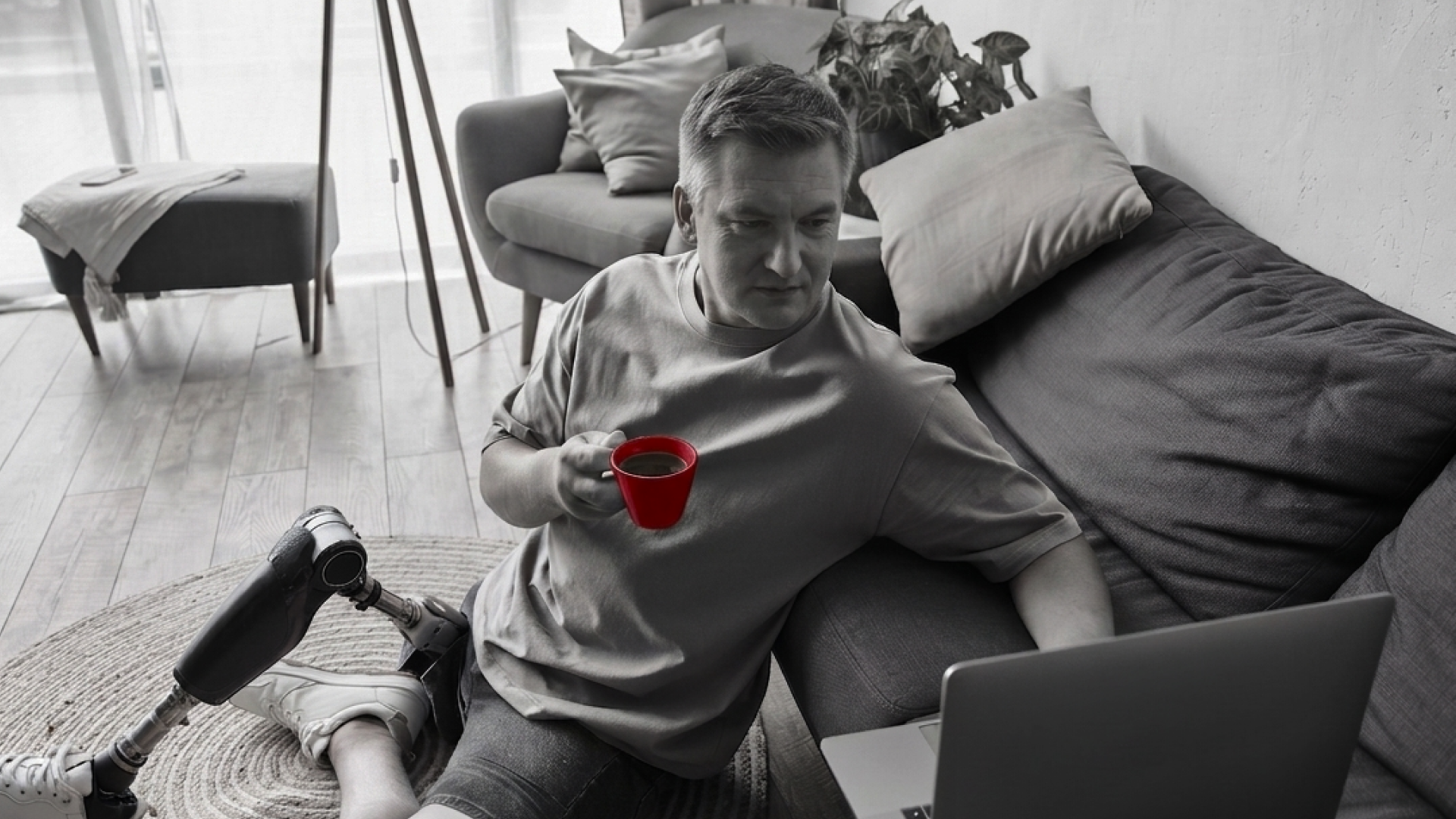 A imagem mostra um homem de meia-idade, sentado no chão, enquanto usa um laptop e segura com uma das mãos uma xícara de café. Ele possui uma prótese de perna.